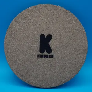 Runder Korkuntersetzer mit dem gravierten Logo von "KINDEKO" in der Mitte. Das Logo besteht aus einem großen, schwarzen Buchstaben "K" und dem Firmennamen "KINDEKO" in stilisierter Schrift darunter. Ideal als stilvolle Tischdeko oder praktisches Accessoire.