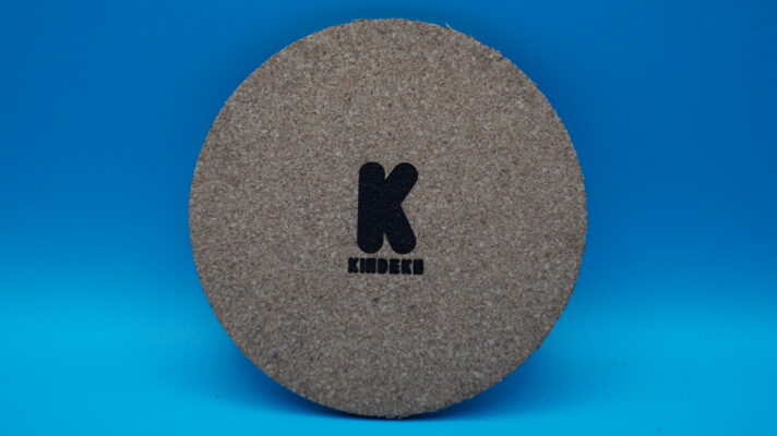 Runder Korkuntersetzer mit dem gravierten Logo von "KINDEKO" in der Mitte. Das Logo besteht aus einem großen, schwarzen Buchstaben "K" und dem Firmennamen "KINDEKO" in stilisierter Schrift darunter. Ideal als stilvolle Tischdeko oder praktisches Accessoire.