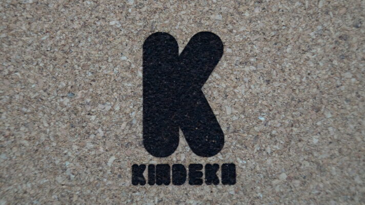 Nahaufnahme eines Korkuntersetzers mit dem gravierten Logo von "KINDEKO". Das Logo besteht aus einem großen, schwarzen Buchstaben "K" und dem Firmennamen "KINDEKO" in stilisierter Schrift darunter.