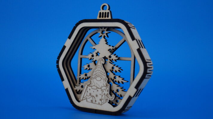 Weihnachtsornament aus Holz mit Tannenbaum, Geschenk aus dem ein Wichtel schaut und mehrschichtigem Rahmen vor einem Fenster.