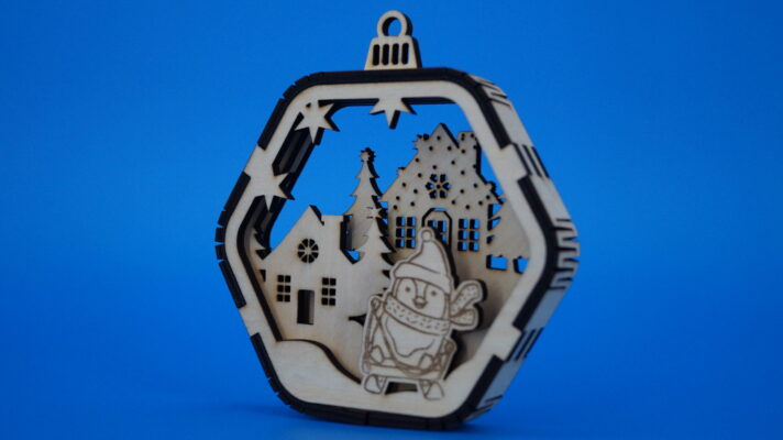 Holz-Weihnachtsornament mit verschneitem Dorf, Tannenbäumen und Schlitten fahrendem Pinguin, mehrlagiger Aufbau