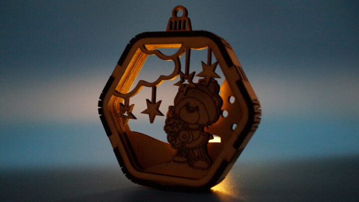 Mehrlagiges Weihnachtsornament aus Holz mit winterlicher Szene, Sternen und Teddy mit Weihnachtsbaum, detailreich gelasert. Von hinten mit einem Teelicht beleuchtet.